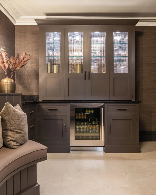 Step inside our moody, speakeasy-inspired bar and media wall project.

This space combines a bespoke Kavanagh bar in rich Hazelwood with a Caesarstone Vanilla Noir worktop, fully integrated appliances including a Quooker tap, Franke sink, and Sub-Zero wine fridge.

The bar’s bi-fold doors reveal a functional serving area with tailored storage for bottles, glassware, and bar tools, while glass shelving set against antiqued bronze mirrors adds a touch of private Soho glamour. Across the room, a Hazelwood media wall with reeded backboards, LED lighting, and a mix of open and closed cabinetry provides a seamless entertainment hub, perfectly blending style and practicality.

Discover the full project and explore every design detail on our blog.